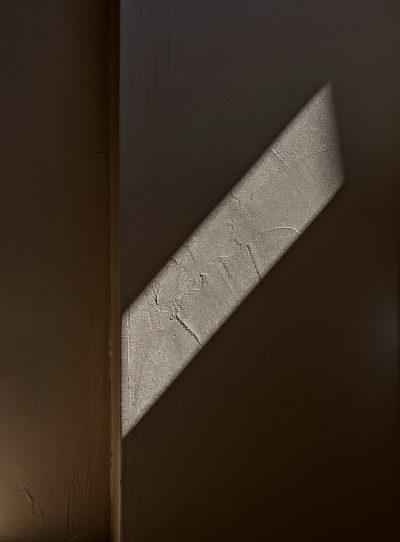 A photo of a wall with an angular shadow cast on it. The light is coming from above and illuminating only half of one side of the room, while the other side has no light. The texture in the shadows looks like concrete or sandstone. --ar 47:64