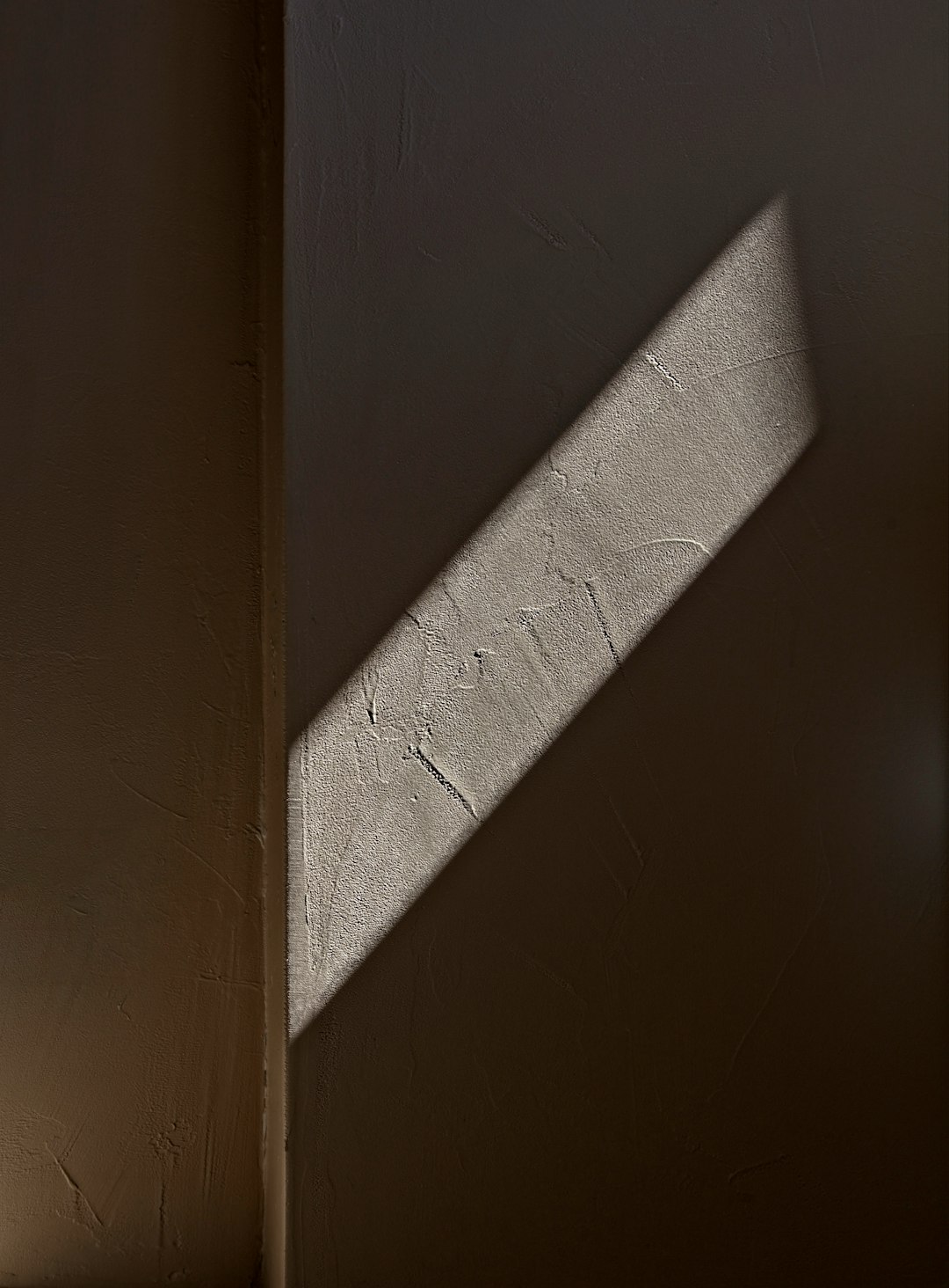 A photo of a wall with an angular shadow cast on it. The light is coming from above and illuminating only half of one side of the room, while the other side has no light. The texture in the shadows looks like concrete or sandstone. –ar 47:64