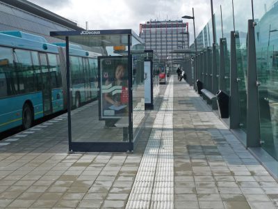 A bus stop with glass walls and transparent signs for advertising on the outside, located at Lautrec square in cushions park of T asteroids near Amsterdam airport, added a big billboard to make it visible from far away. The street is very wide and has large sidewalks. There is also a public toilet next door. In front there should be empty space for waiting, with a concrete floor, grey tiles and concrete stairs. The photo was taken in the style of Canon eos r3. --ar 4:3