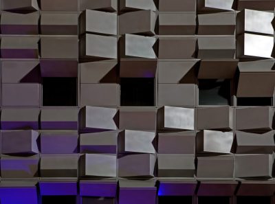 A photograph of an abstract building facade made from shiny metal cubes, reflecting light and creating a dynamic pattern in shades of gray, purple, and blue. The focus is on the intricate arrangement of these cubes forming a modern architectural structure with geometric precision. Soft lighting highlights their reflective surfaces against a dark background. --ar 128:95