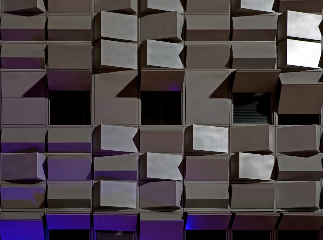 A photograph of an abstract building facade made from shiny metal cubes, reflecting light and creating a dynamic pattern in shades of gray, purple, and blue. The focus is on the intricate arrangement of these cubes forming a modern architectural structure with geometric precision. Soft lighting highlights their reflective surfaces against a dark background. –ar 128:95