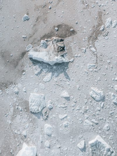 flat lay top view of the antarctic ice shelf with small rocks and debris scattered around, no snow on ground, no people or animals visible, drone shot, overhead view, white color palette, natural lighting, hyper realistic --ar 95:128