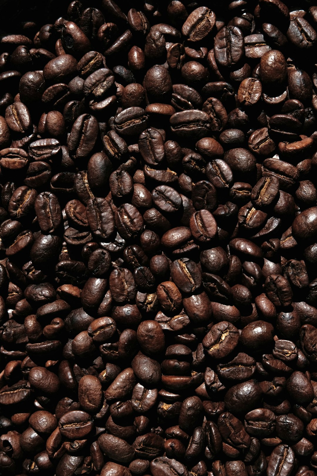 A pile of dark coffee beans. A photo taken from above. The background is black, with the texture of fresh roasted beans visible on top. It’s a closeup shot. This scene creates an atmosphere that emphasizes quality and naturalness in product presentation. The overall composition exudes freshness, vitality, warmth, depth, and high definition. High resolution photography captures every detail in the style of a professional photographer. –ar 85:128