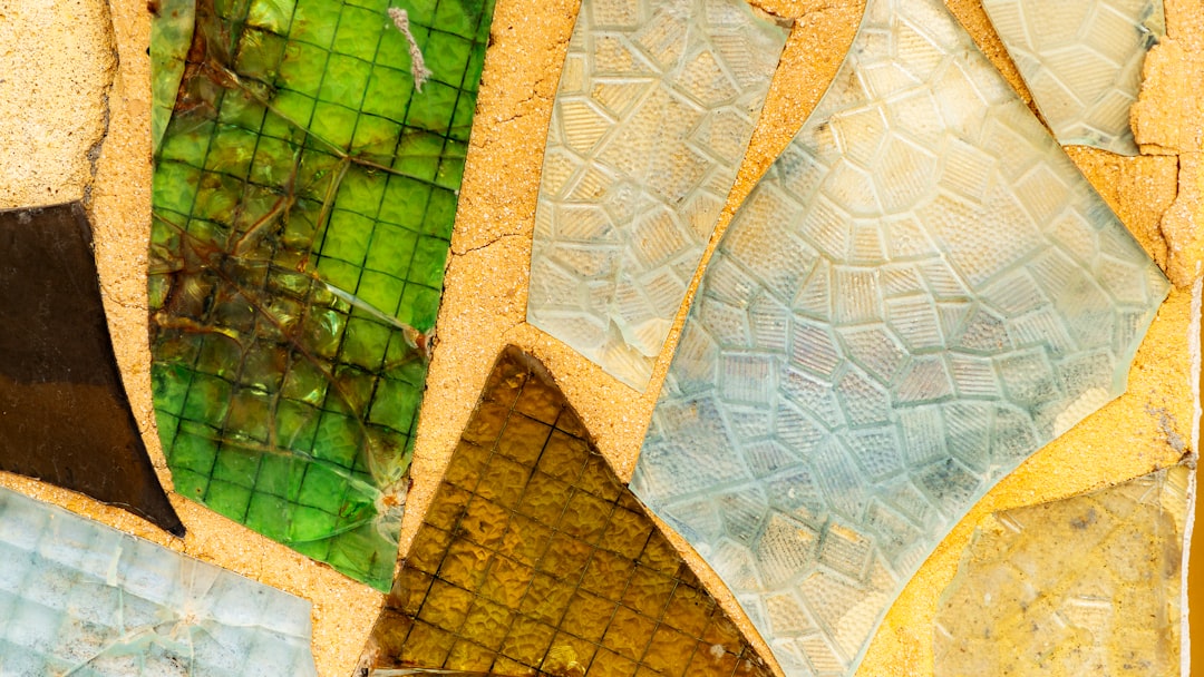 Close up of a colorful and textured glass mosaic featuring green, yellow, brown, blue and white patterns in the style of Antoni Gaudi’s architecture. The abstract patterns appear broken and cracked against a flat background with intricate and detailed designs. Natural lighting brings out the beautiful, stunning details of the mosaic. –ar 16:9