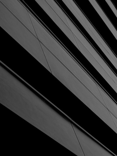 Black and white photography of diagonal lines on the side of an office building, creating geometric patterns with sharp angles. The background is a dark grey to accentuate the details in black, providing contrast that adds depth and texture to the composition.