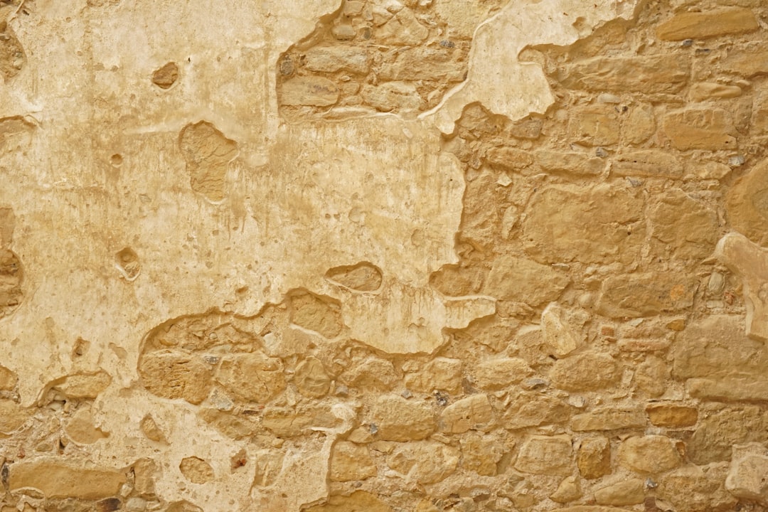 An old wall of yellowish brown stone, with rough and visible textures that give it an aged appearance. The background is solid and neutral in color to highlight the detailed texture of the stonework. This backdrop would be suitable for various design projects or as part of artistic illustrations. It could create an atmosphere reminiscent of ancient structures or historical architecture in the style of various artists. –ar 128:85