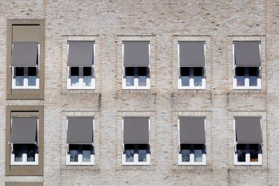 A closeup of the exterior wall of an apartment building with multiple windows, each window is covered in gray shades blinds. The building's facade is made from light brown brick and has neutral tones. In some rooms you can see people sitting at their desks in the style of them working on computers. --ar 128:85