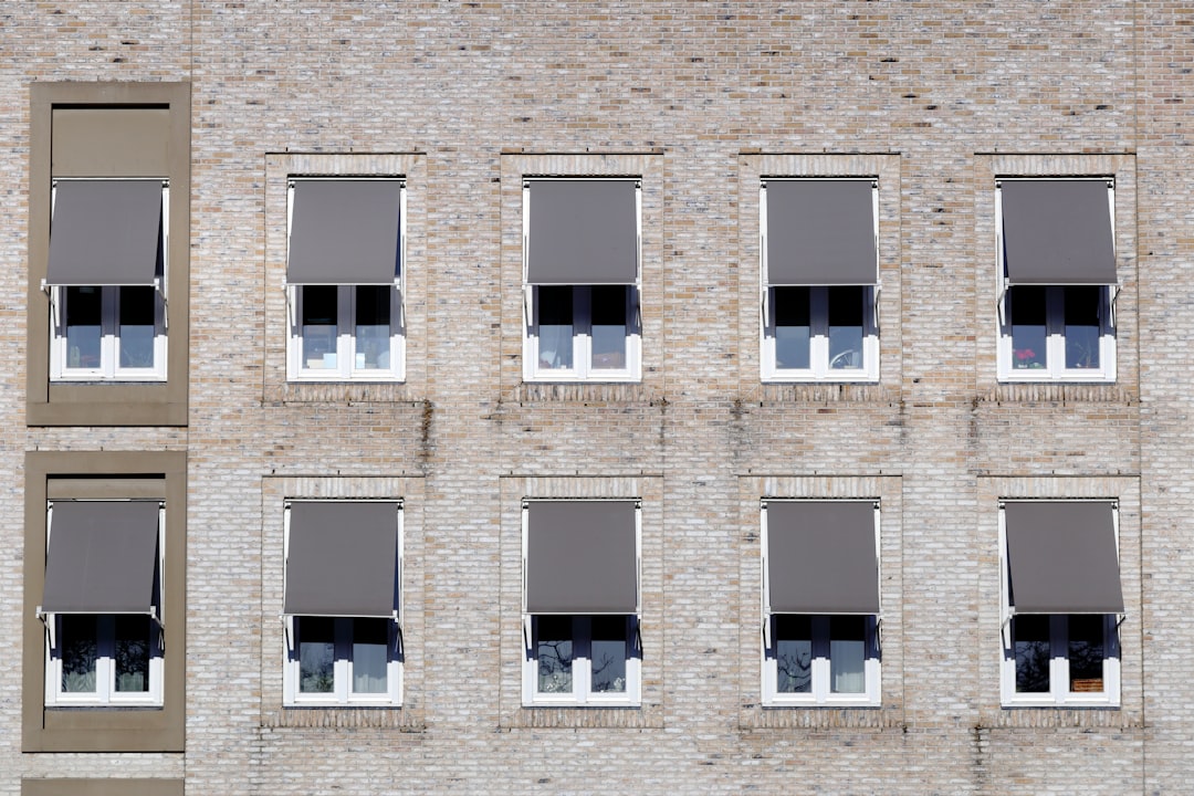 A closeup of the exterior wall of an apartment building with multiple windows, each window is covered in gray shades blinds. The building’s facade is made from light brown brick and has neutral tones. In some rooms you can see people sitting at their desks in the style of them working on computers. –ar 128:85