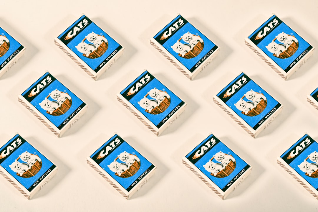 A grid of white match boxes with the word “CATS” printed on them, each containing one blue and brown cat-shaped cigarette match, arranged in rows against a pure background. The illustrations feature cats depicted as pet toys or accessories, showcasing playful poses, and include subtle details like fur textures to enhance realism. This design is for an advertising campaign that celebrates casual moments between friends and their feline companions, in the style of playful pet illustrations. –ar 128:85