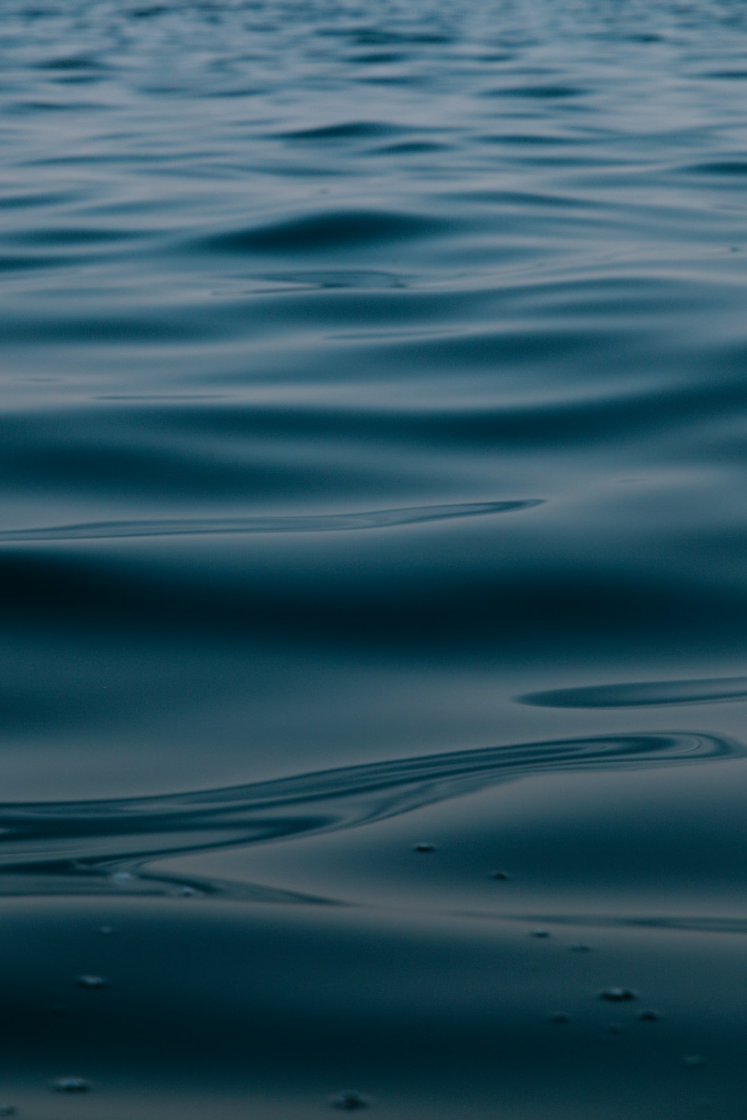 A closeup of the calm, dark blue water surface with gentle ripples in the background. The focus is on the subtle waves and reflections that create an abstract pattern. This scene evokes tranquility and harmony, symbolizing stillness and peace. Shot in the style of Canon EOS R5 at F2, ISO400, Shutter Speed 360, with 8k resolution, hyper realistic, cinematic light, and very detailed from a high angle shot. –ar 85:128