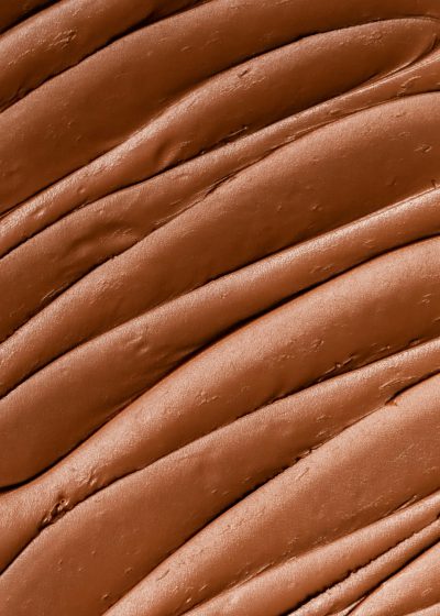 A closeup of the texture and color of liquid foundation in a chocolate brown color. The skin is slightly wavy with natural patterns, creating an elegant appearance. A macro shot focuses on one area to highlight details such as fine lines or delicate textures. This image should convey luxury and beauty through its detailed depiction of smooth skin in the style of elegant appearance. --ar 91:128