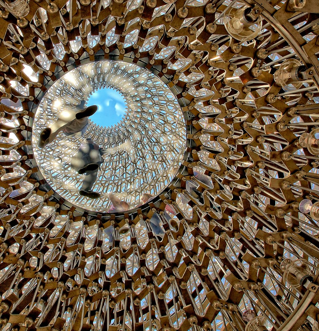 Aerial view of the ceiling of an art installation made from thousands of cutlery pieces arranged in intricate patterns, creating a surreal and eyecatching visual effect with reflections on the shiny surfaces, a clear blue sky visible through one section of glass, Photography, shot using a wideangle lens to capture all elements of the dome, –ar 123:128