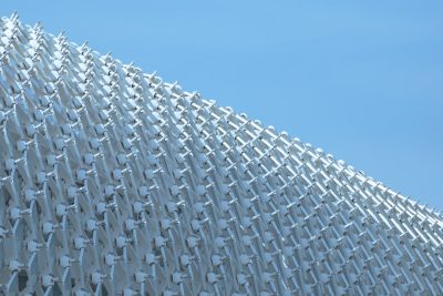 A large white metal building was covered in hundreds of pale blue and off-white plastic shapes that resembled tiny chairs arranged to create an undulating wall, creating the impression of one very tall chair covering it all. The background was a clear sky, with a close up, low angle photograph taken in the hyper realistic style. --ar 128:85