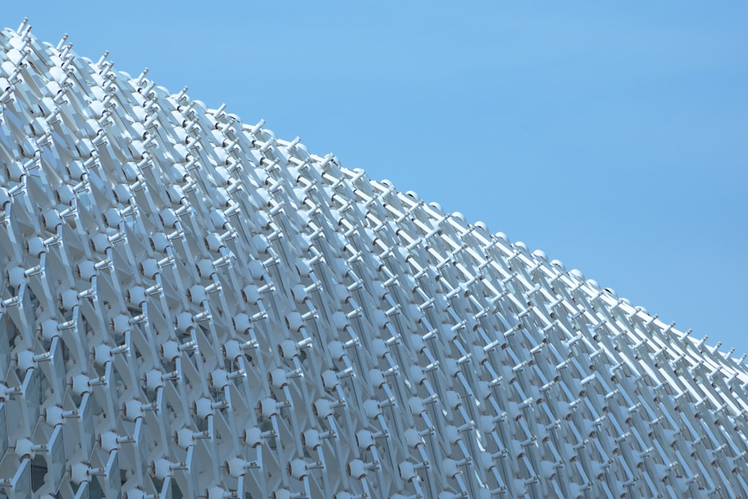 A large white metal building was covered in hundreds of pale blue and off-white plastic shapes that resembled tiny chairs arranged to create an undulating wall, creating the impression of one very tall chair covering it all. The background was a clear sky, with a close up, low angle photograph taken in the hyper realistic style. –ar 128:85