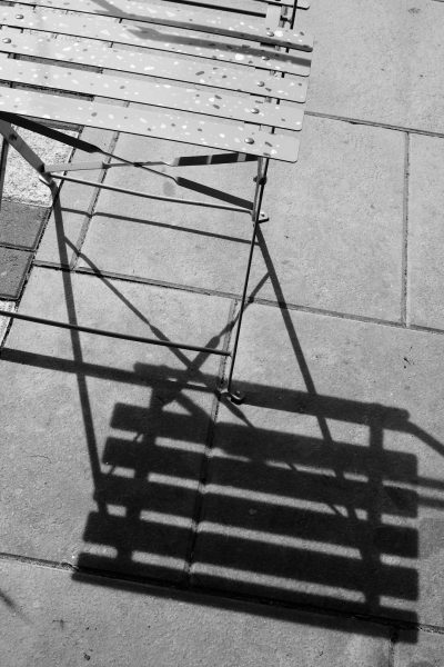 A black and white photo of a shadow from an outdoor cafe table on the ground, with a metal frame chair next to it. The shadow and chair are in the style of [Fan Ho](https://goo.gl/search?artist%20Fan%20Ho). --ar 85:128