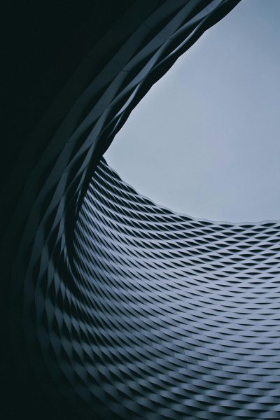 Minimalist photography, award winning, a large dark grey metal structure with undulating lines and subtle patterns on the surface, looking up at it from below, the sky is blue, clean background, low angle shot, cinematic, grainy film in the style of. --ar 85:128