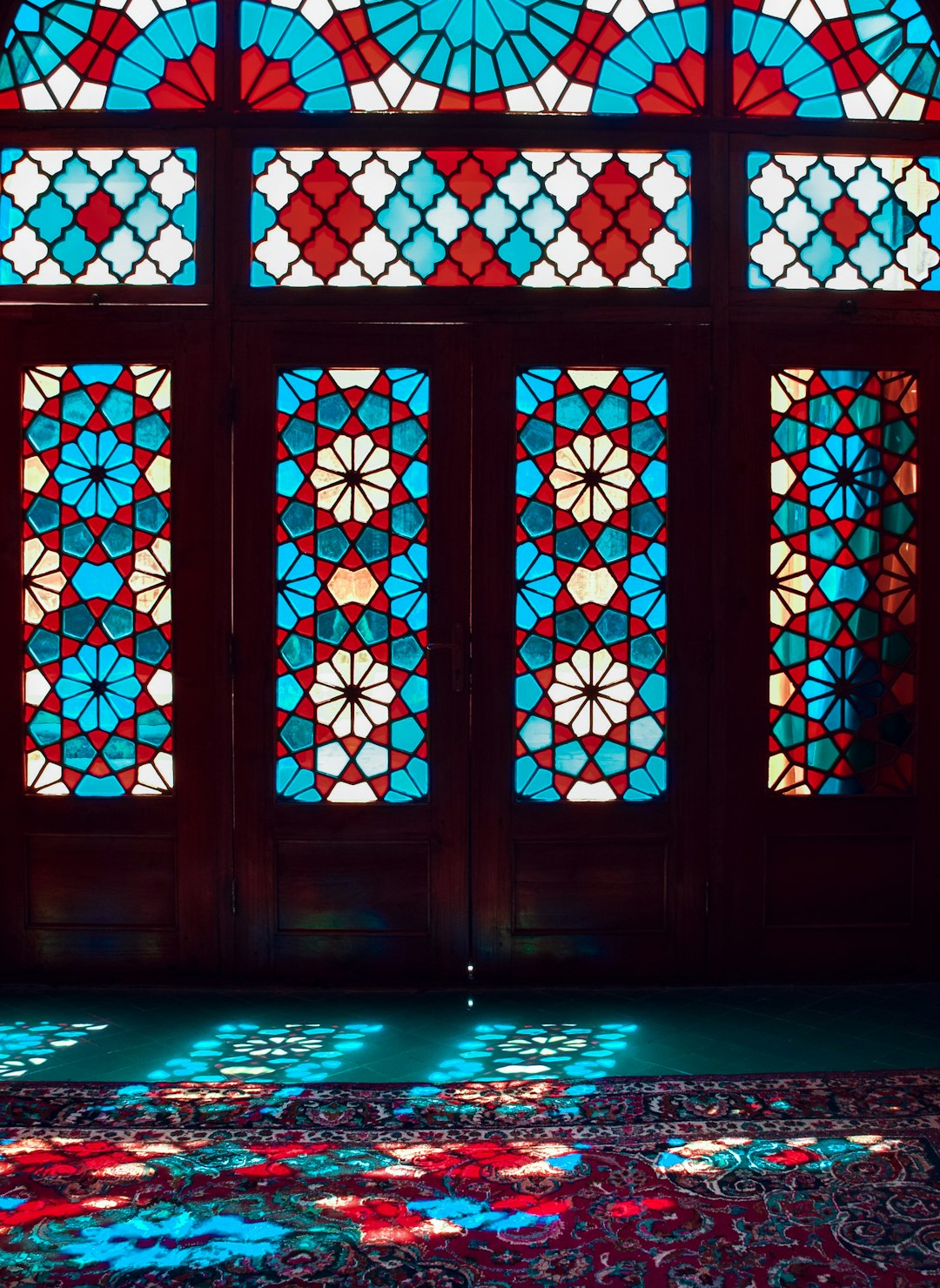 an intricate geometric patterned glass window in an old persian house, colorful light shining through the windows onto Persian carpet on floor below, red and blue hues, beautiful –ar 93:128