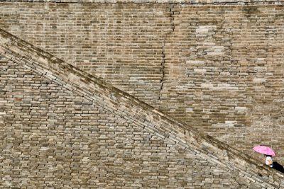 A photograph of an ancient Chinese brick wall with one person walking up the stairs in a top view style, carrying a pink umbrella, minimally editing the original text and removing any Chinese characters. The photograph shows one person walking up the stairs in the style of a top view, carrying a pink umbrella. --ar 128:85