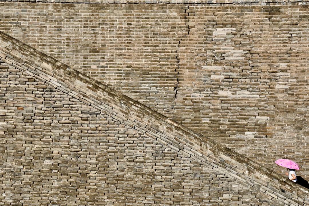 A photograph of an ancient Chinese brick wall with one person walking up the stairs in a top view style, carrying a pink umbrella, minimally editing the original text and removing any Chinese characters. The photograph shows one person walking up the stairs in the style of a top view, carrying a pink umbrella. –ar 128:85