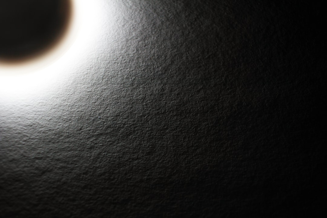 close up of black matte surface, small smooth circular patch in the middle with light shining on it, shot from above, low angle, macro photography, minimalism, monochrome, texture focus, depth of field, shadow play, backlit –ar 128:85