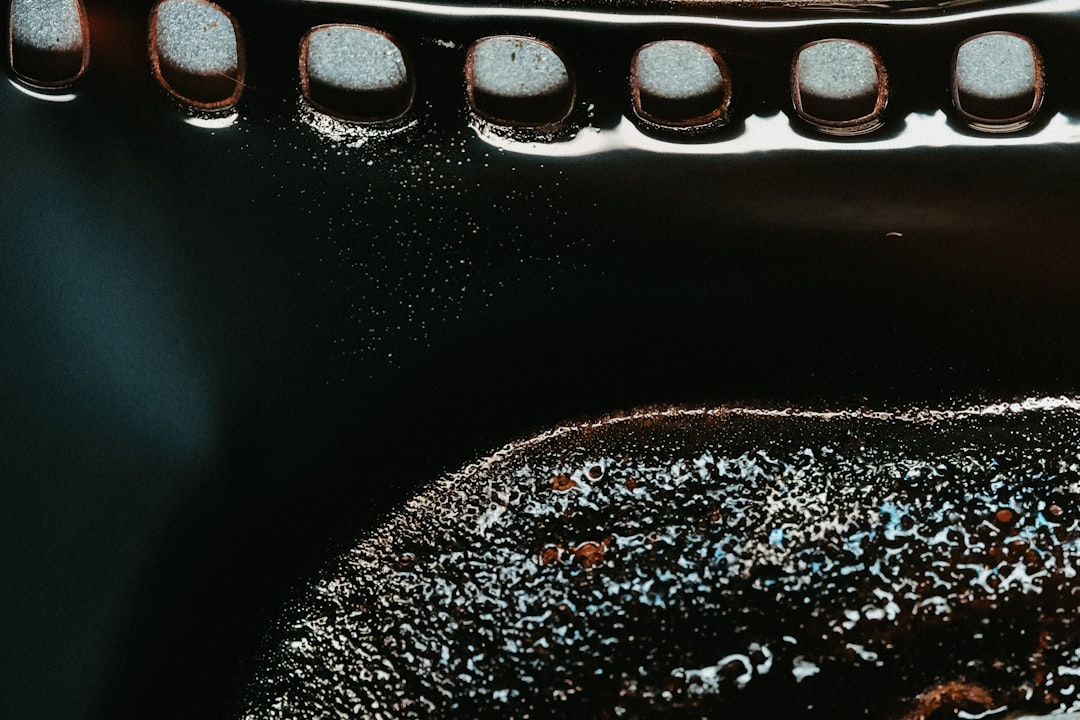 A closeup of the inside bottom edge of an iron cast pan, filled with oil and black coffee powder. The camera angle is from above at eye level, focusing on details like metal textures and small bubbles. It’s a dark room with a soft light source highlighting these features. The image is in the style of gas flame. –ar 128:85