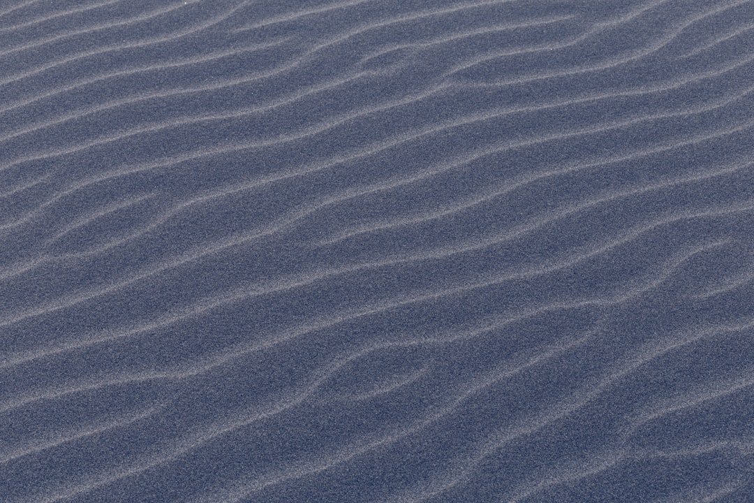 A high-resolution texture of sand ripples, captured from above in the desert at dusk. The textured surface is navy blue with subtle gray patterns and lines that create an abstract pattern reminiscent of ocean waves. It is a tranquil yet mysterious background for design projects. –ar 128:85