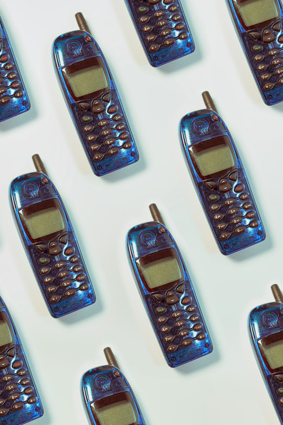 A pattern of blue cell phones, all lying on their side, against a white background. The cell phones each have an illuminated screen and are made from plastic with visible textures. Each one shows the same stock photo website interface. –ar 85:128