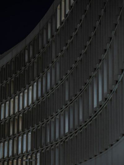 Night photo of the curved facade wall with vertical and horizontal lines on the concrete panels. Soft light illuminates the architectural photography with a building closeup from a low angle shot, cityscape in the background providing an urban atmosphere. The minimalistic style features subtle lighting and a high resolution sharp focus showing depth of field and detailed textures of the architectural details in the style of an architectural masterpiece. --ar 3:4