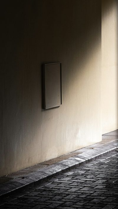 A photo of an empty picture frame on the wall, hanging at eye level on a cobblestone street in Rome, taken from a side angle. The frame is blank and black with no text or graphics inside it. It is illuminated by soft natural light coming through the window above, casting gentle shadows across its surface. A shadow falls to one side, adding depth to the scene. --ar 9:16