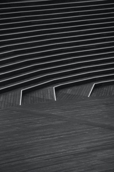 A black and white photograph of an abstract, geometric composition featuring curved lines on the floor leading up to steps with visible wood textures. The background is a monochromatic gradient that transitions from dark at the top to light towards the bottom, creating a sense of depth in perspective. --ar 85:128