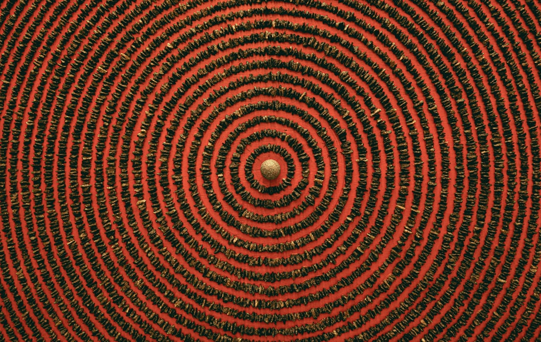 A red circular pattern with black lines, creating an intricate and symmetrical design on the fabric surface. The background is dark brown, adding depth to the composition. A small gold ball in the center of each circle adds contrast and detail. This image captures macro photography aesthetics by capturing every line and texture. It’s a detailed closeup shot that highlights both fine print quality and the overall aesthetic appeal of materials. –ar 8:5