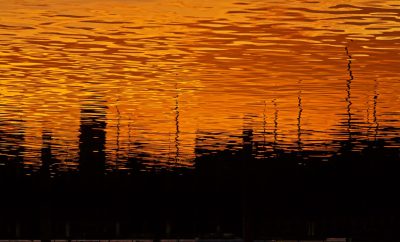 The reflection of the orange sunset in water ripples on an abstract background. The silhouette is of trees and buildings, creating a contrast between lightness and darkness. --ar 128:77