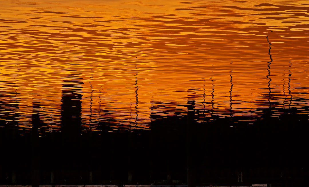 The reflection of the orange sunset in water ripples on an abstract background. The silhouette is of trees and buildings, creating a contrast between lightness and darkness. –ar 128:77