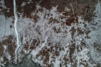Aerial view of an abstract landscape with brown and white patterns, ice on the ground, a blue river in between, top down perspective, shot from above, photography. The landscape shows patterns in the style of ice on the ground and a blue river between, from a top down perspective as if shot from above, in the photography style.