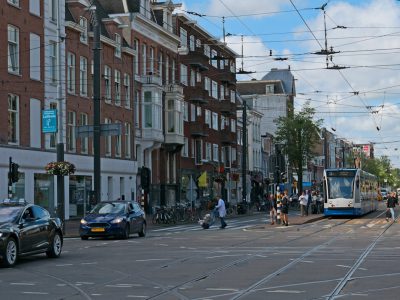 A photo of an Amsterdam street with electric trams, cars and people walking on both sides of busy streets in Amsterdam on a summer day with a blue sky. The photorealistic landscape shows street scenes with many buildings and car traffic, with people crossing an intersection or sitting at outdoor chairs near shops. The photo was taken in the style of Canon EOS. --ar 4:3