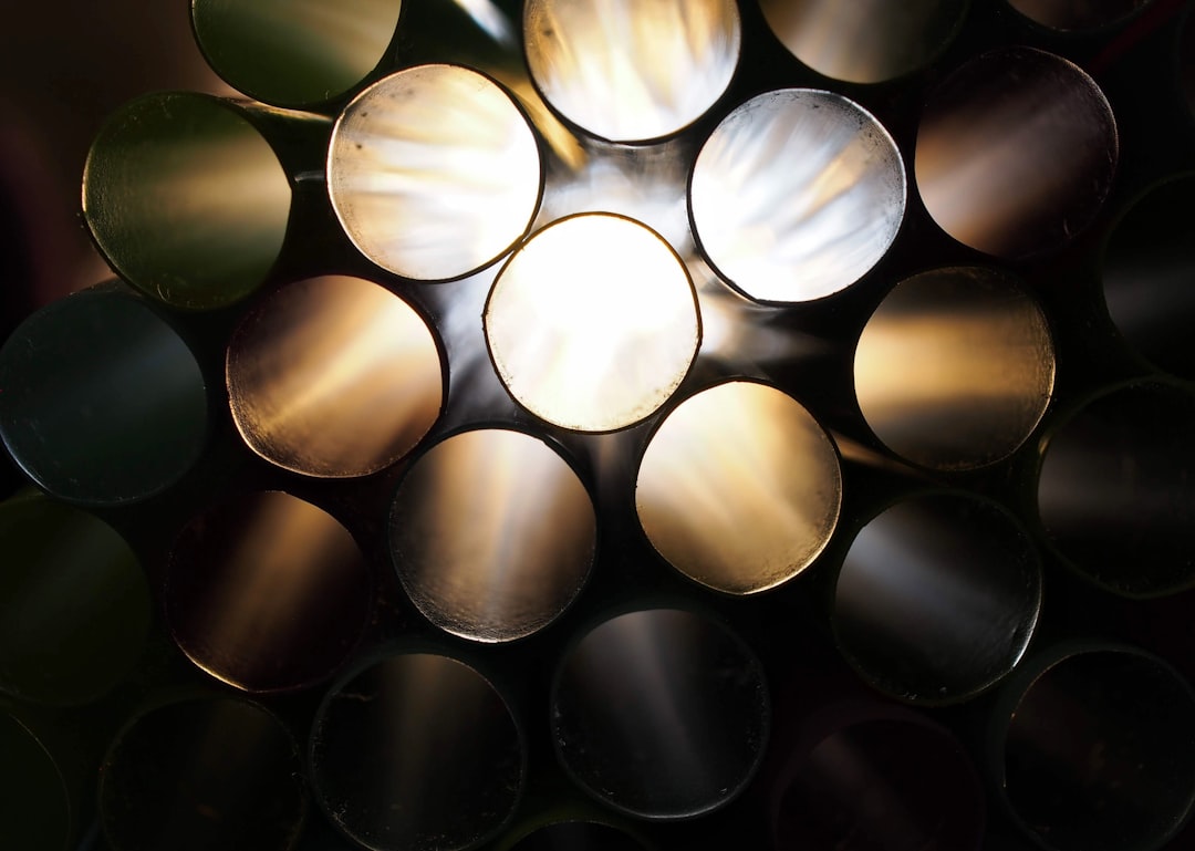 A close up of an array of round metal pipes, lit from above by the sun in a dark room. The light is coming through the holes and illuminating them with a warm glow. In front there is one pipe that has been painted white. It reflects some of the other colors in the style of the artist. –ar 128:91