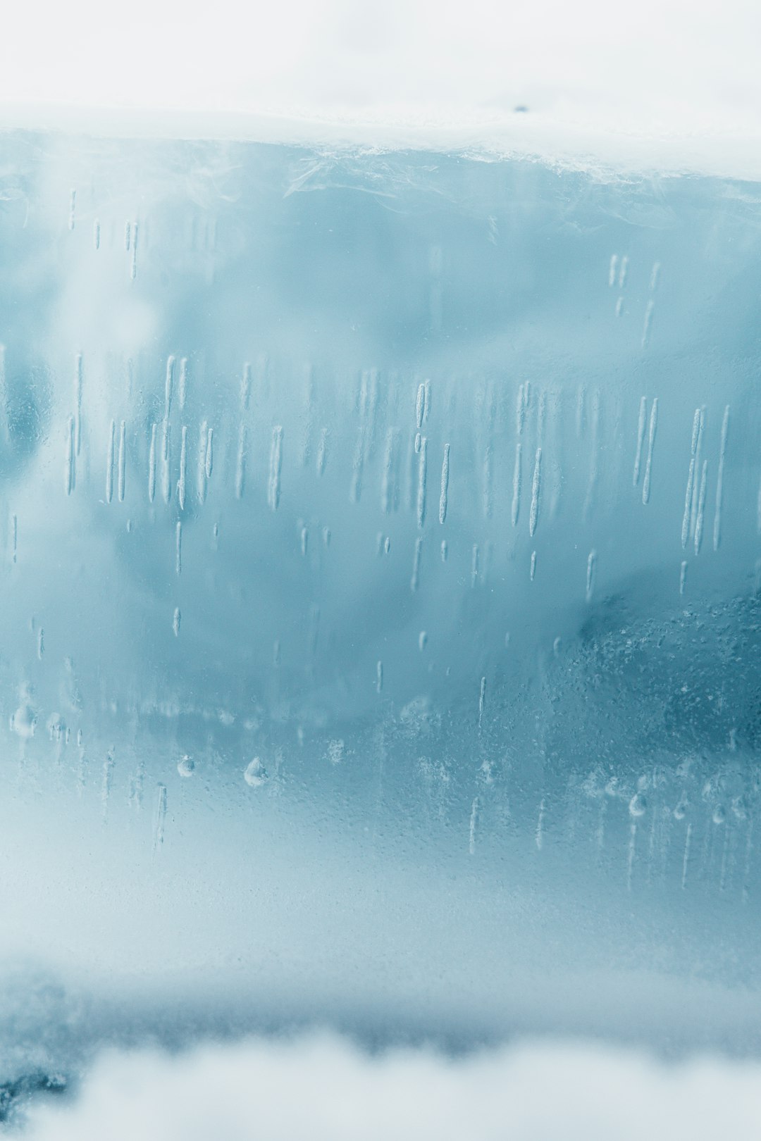 A photo of a close-up shot of an ice wall, covered in raindrops, with soft blue and white tones, a foggy background, taken with a Canon EOS R5 at F2, ISO30, and 89MM. Soft lighting creates a misty, atmospheric, and ethereal dreamlike style. –ar 85:128