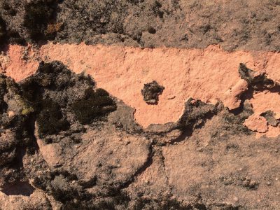 a red clay wall with black lichen on it, the left side of which has an orange line drawn in. The drawing is made by hand and appears to have been done using oil paint or some similar medium. There's no text on that line but there seems like another small dot or three along its length. This photo was taken from above so we can see only half of one edge of the red wall and part of the other. It must be a real photograph as I am pretty sure you would not make up something like this for fun, and your signature could stand out well against the wall. --ar 4:3