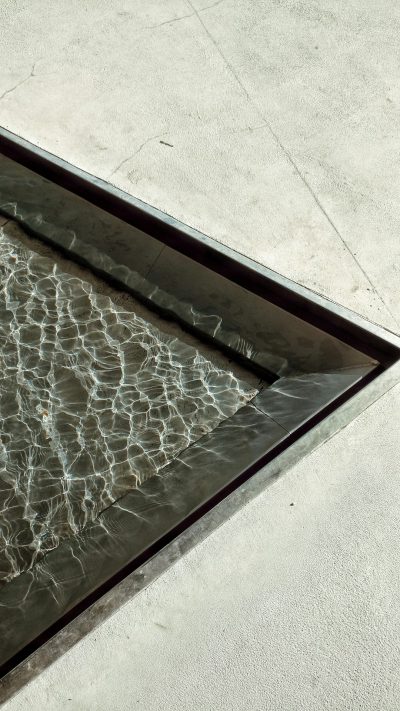 An overhead shot of the edge and corner detail on an elegant, flat surface with water flowing through it. The focus is on capturing the reflection in the glass as well as showcasing its sleek design. This perspective highlights the contrast between the light concrete floor and dark metal frame. --ar 9:16