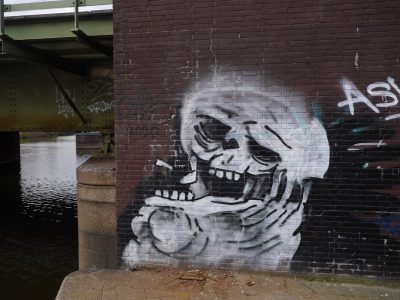 Graffiti of a laughing skull smoking on the wall next to a river, under a bridge in Amsterdam in the style of [Banksy](https://goo.gl/search?artist%20Banksy). --ar 4:3