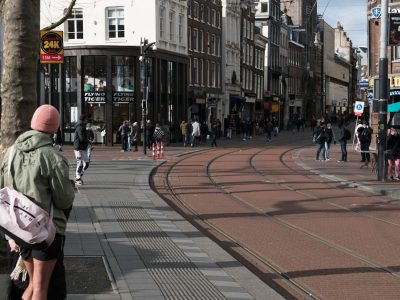 Photo of a busy Dutch street with people. A wide angle shot shows one person standing on the left side wearing a light green jacket and pink hat looking to their right. In front, there is an empty square where two thin brown tram tracks can be seen running straight through it. There should be some pedestrians walking towards the camera but more going away from it. The sign "24K" is visible at the top corner of the picture. Buildings can be seen in the background on a sunny day. --ar 128:95