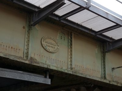 A steel bridge with an engraved "K momSteamer" emblem on the side, seen from below. The structure is aged and rusted, but it's still functional for passing vehicles. There’s visible wear in its metalwork, indicating use over time. In the background there’s another part of that same iron frame showing signs of age and dis poems about bridges, chrome, minimalistic --ar 4:3