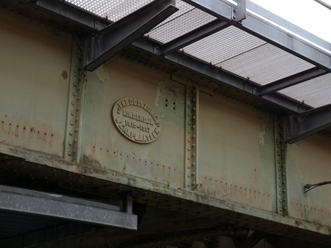 A steel bridge with an engraved “K momSteamer” emblem on the side, seen from below. The structure is aged and rusted, but it’s still functional for passing vehicles. There’s visible wear in its metalwork, indicating use over time. In the background there’s another part of that same iron frame showing signs of age and dis poems about bridges, chrome, minimalistic –ar 4:3