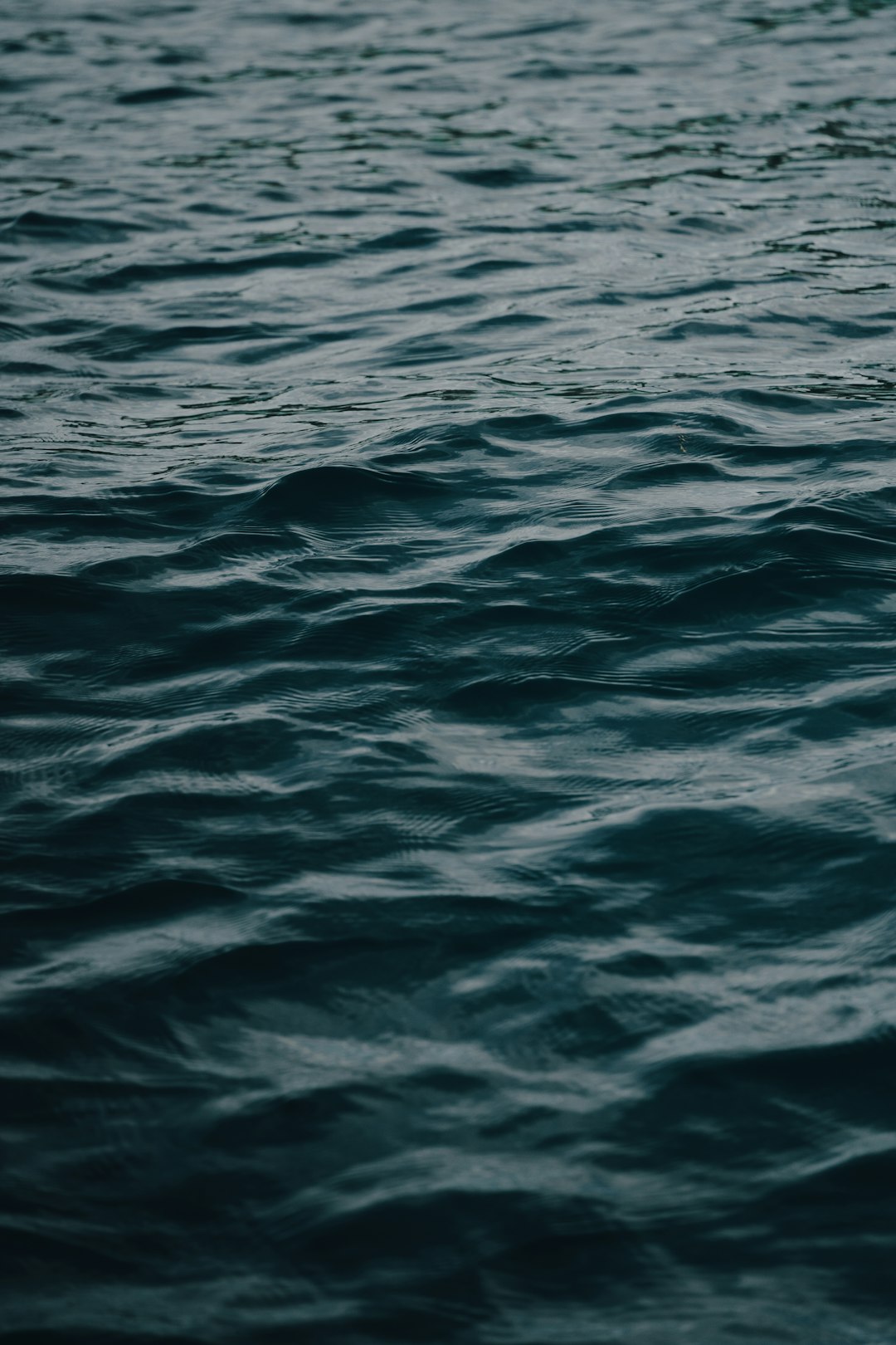 Dark blue water, flat sea surface, shot from the side, minimalist style, high definition photography, real life scenes, closeup shots, blurred focus, and no waves on the water’s surface. The entire scene is dark gray with subtle color changes in shades of deep green and navy blue. A calm atmosphere envelops everything. This photo was taken in the style of Sony Alpha A7 III camera with an aperture setting of f/8, ISO 200, focal length of 56mm, and shutter speed of one using multiple exposure mode. –ar 85:128