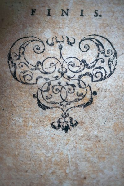 The word "FINIS." written in an ornate, antique script on aged parchment paper with intricate ink designs and patterns around the text. The letters have been carved into old parchment material in the style of handwriting. There is a large heart-shaped emblem at its center, symbolizing love or romance. In soft lighting, the details of each letter and design can be clearly seen. This artwork has elements reminiscent of medieval manuscript art, evoking nostalgia for historical illustrations. --ar 85:128