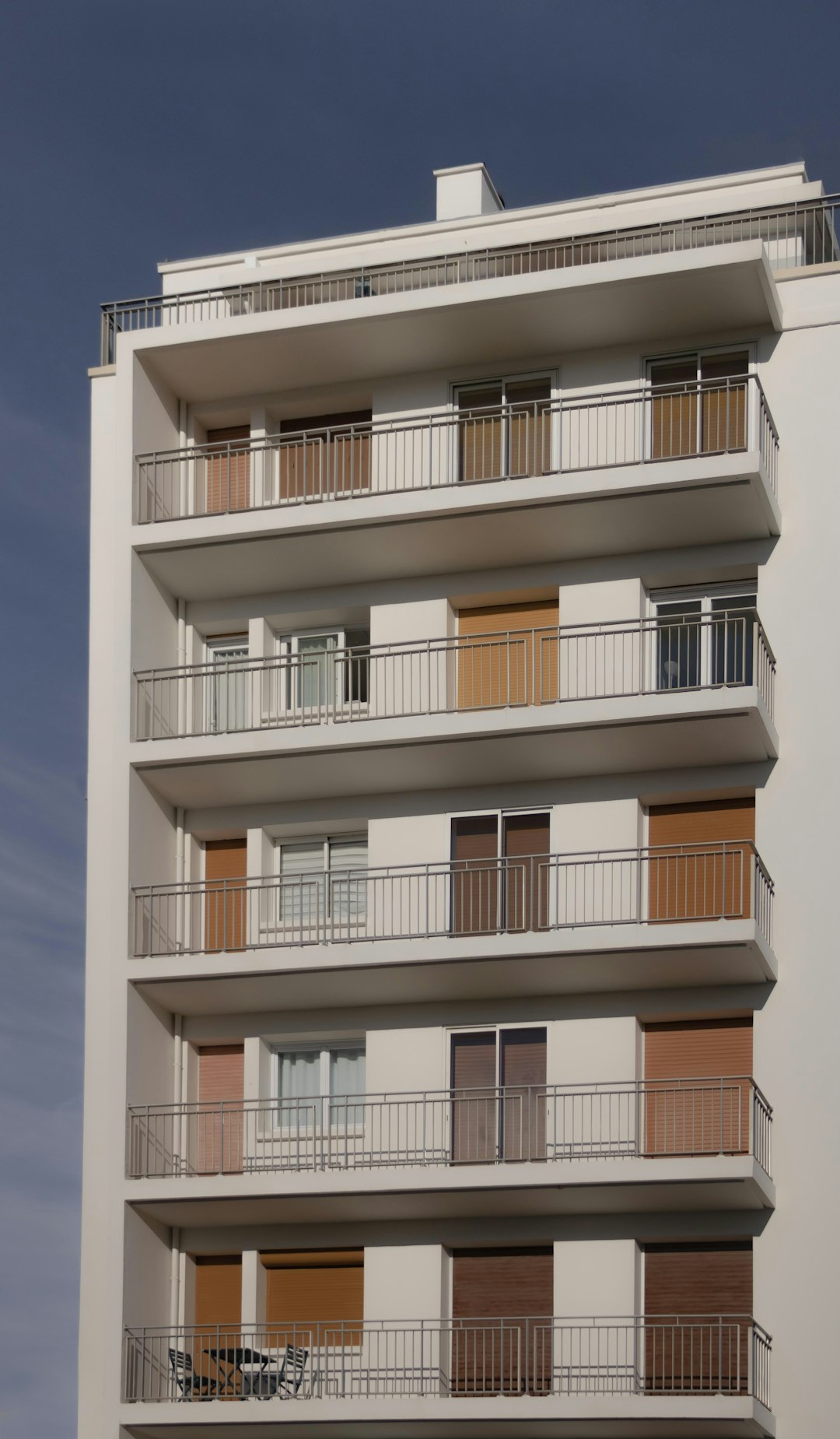 A clean white modern building with balconies, with wooden shutters on the balcony doors, with one floor slightly higher than other floors, in shades of brown and beige colors, against a dark blue sky, photo realistic, taken in the style of Canon EOS. –ar 37:64