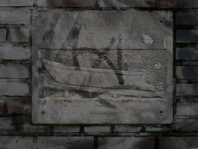 A stone slab with the inscription "Kangonden" and an engraving of small boat on it, is mounted against a gray brick wall. The text reads in Dutch 'ik kijkt op de watermerk van brood macrophotography --ar 4:3