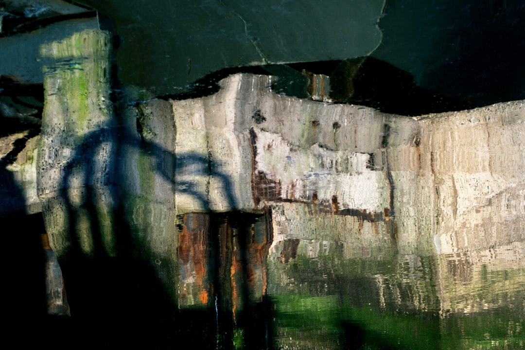 A closeup of the shadow cast in the style of an old, weathered wall on water, with reflections and shadows creating intricate patterns in shades of green, brown, grey, and white. The background is blurred to focus attention on the textures and colors of concrete, water ripples, and faint outlines of other structures. –ar 128:85