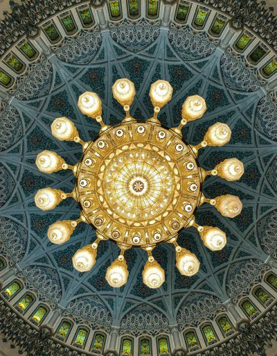 The grand chandelier of the Omani National Palace was built in the style of Sultan Q. It comprehensively includes many golden light bulbs, surrounded by ornate geometric patterns in green and blue colours with intricate details. The photo was taken from below. --ar 99:128