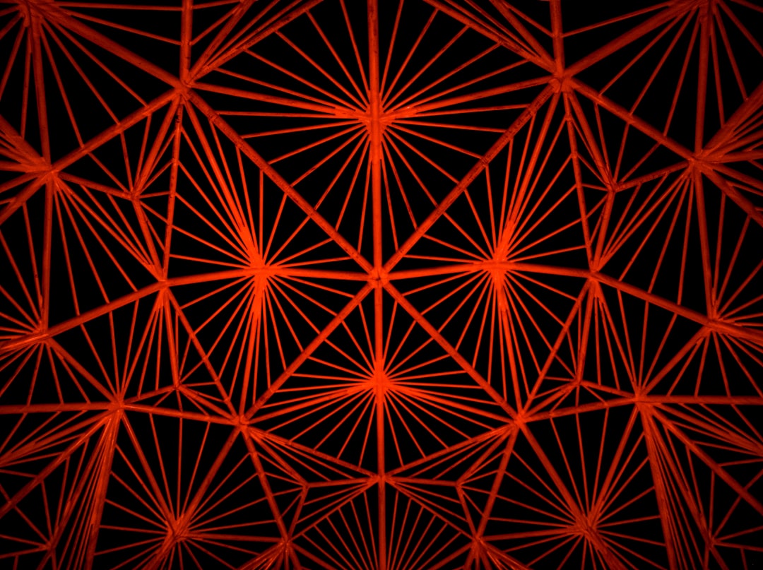 Red and black background with a geometric pattern of red lines, symmetrical composition, and interlacing lines forming an intricate network of triangles. The pattern conveys depth and perspective in the dark space. –ar 128:95